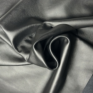 May include: Close-up of a sheet of black leather with a textured surface. The leather is folded and draped, creating shadows and highlights that emphasize its supple quality. The material appears smooth and durable.