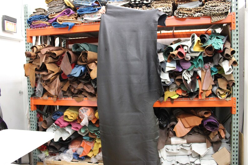 K&ouml;nnte beinhalten: Ein gro&szlig;es Regal gef&uuml;llt mit Lederrollen in verschiedenen Farben, darunter Braun, Beige, Schwarz, Gr&uuml;n und Blau. Das Leder ist gestapelt und gerollt, und einige St&uuml;cke h&auml;ngen an den Regalen.