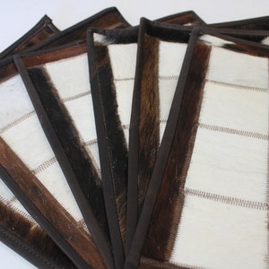 May include: Set of six brown and white cowhide placemats with a stitched border. The placemats are made of natural cowhide and have a rustic, farmhouse style.