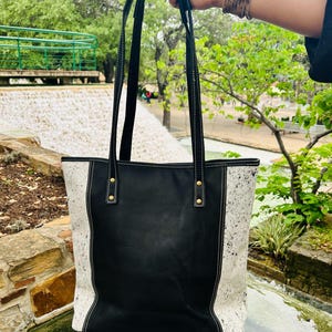 May include: Black leather tote bag with white cowhide accents and gold hardware. The bag has two top handles and a white stitch detail.