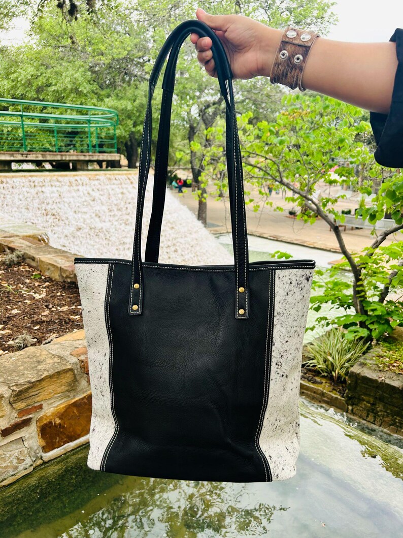 May include: Black leather tote bag with white cowhide accents and gold hardware. The bag has two top handles and a white stitch detail.