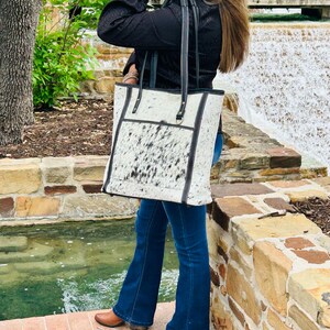 May include: A white and black cowhide tote bag with black leather straps and trim. The bag has a zipper closure and a large pocket on the front.