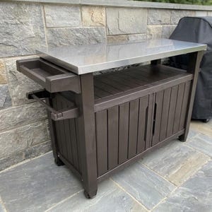 Op de afbeelding: Een donkerbruin buiten-voorbereidingsstation met een rechthoekig roestvrijstalen werkblad. De unit bevat een onderste opbergkast met verticale latten, een handgreep aan de zijkant en een klein bijgevoegd dienblad. Het staat op vier wielen.