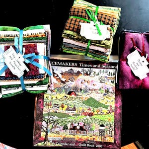May include: Three fabric bundles tied with ribbons and white tags labeled Sky, Water and mountain bundle, Green bundle, and Border and binding bundle, arranged around a Peacemakers Times and Seasons A Calendar and Quilt Book 2000.