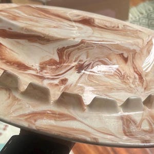 May include: Oval ashtray with a cream and brown marbled design. The ashtray has a series of notched rests for cigarettes and cigars. The ceramic ashtray is a vintage piece.