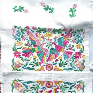 Könnte beinhalten: Weißes besticktes Stoffstück mit farbenfrohen Vogel- und Blumenmustern. Die Stickerei verwendet rosa, gelbe, blaue und grüne Fäden. Der Stoff zeigt ein sich wiederholendes Design von Vögeln und Blumen, ideal für die Heimdekoration.
