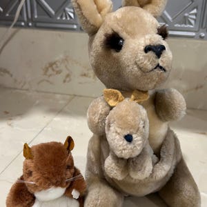 May include: A plush kangaroo toy with a joey in its pouch, and a small brown and white hamster toy. The kangaroo is tan with dark eyes and a small bow tie. The hamster has a white belly and long whiskers.