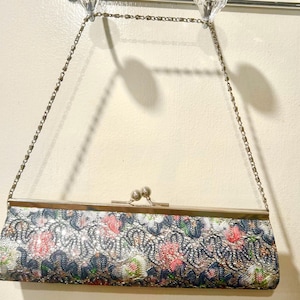 May include: A vintage clutch purse with a silver chain strap. The rectangular purse features a floral pattern in pink, green, and blue on a silver background. The clasp is silver, and the chain is delicate.