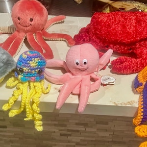 May include: A collection of plush sea creatures, including a gray stingray, a red octopus, a pink octopus, a red crab, a blue and orange crocheted octopus, and a crocheted jellyfish with yellow tentacles.