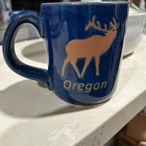 Könnte beinhalten: Eine blaue Keramik-Tasse mit Henkel. Die Tasse zeigt eine goldfarbene Elch-Silhouette und das Wort "Oregon" in weißer Schrift. Die Tasse steht auf einer weißen Oberfläche.