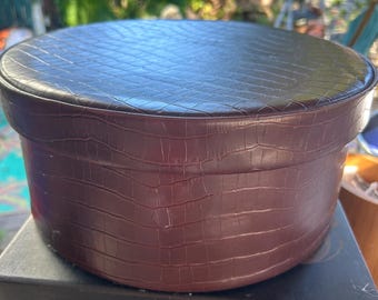May include: A round, dark brown storage box with a textured, embossed crocodile skin pattern. The container features a matching lid that fits securely over the top, creating a cylindrical shape with a consistent, deep brown color throughout.