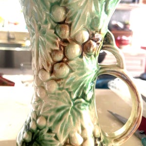 May include: A ceramic pitcher featuring a raised, three-dimensional relief pattern of grape clusters and vine leaves. The surface is finished with a glossy glaze in shades of light green, cream, and brown accents, with a curved handle on the side.