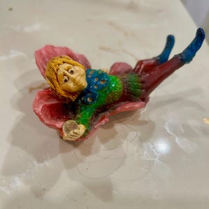 May include: A small figurine of a person with blonde hair lying on a large pink flower petal. The figure wears a green top with blue accents, dark red trousers, and blue boots. The person is holding a small, clear, circular object in one hand.