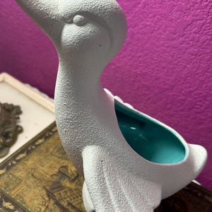 Könnte beinhalten: Ein weißer Keramik-Enten-Pflanztopf mit strukturierter Oberfläche. Der Innenraum ist türkis. Die Ente hat einen langen Hals und einen nach oben zeigenden Schnabel. Der Pflanztopf steht auf einem Vintage-Buch.
