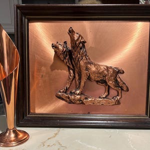 May include: A copper-colored vase with a flared top and a round base stands next to a framed copper relief. The relief depicts two howling wolves on a copper background, framed in dark wood.