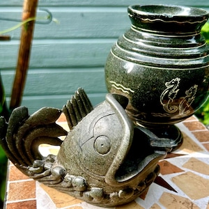 May include: A dark green stone ashtray in the shape of a fish with an open mouth, next to a dark green vase with seahorse designs. The vase has horizontal lines and a wide opening. Both items are on a mosaic tile surface.