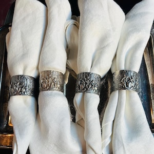 May include: Four white linen napkins are held by silver-coloured napkin rings. Each ring features a detailed relief design of figures in a classical setting. The napkins are neatly folded, creating a formal and elegant presentation, suitable for a dining table setting.