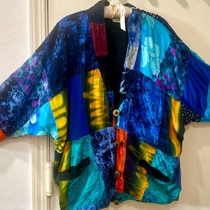 May include: A patchwork jacket in shades of blue, teal, yellow, and orange. The jacket features a variety of fabric patterns and textures, with a button-up front and two pockets. The sleeves are wide and the collar is black.