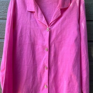 May include: A bright pink button-down shirt with long sleeves. The shirt has a classic collar and is made of a textured fabric. The buttons are a translucent pink. The shirt is hanging on a floral patterned hanger.