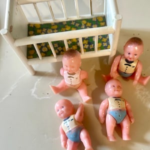 May include: A vintage white wooden doll crib with a floral patterned mattress, accompanied by four small plastic baby dolls. The dolls are pink with painted features, wearing blue diapers and cream-colored vests.