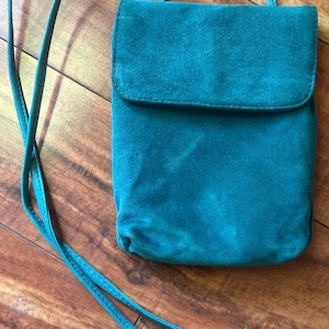 May include: A teal-colored crossbody bag with a rectangular body and a fold-over flap closure. The bag features a long, thin matching shoulder strap and is constructed from a soft, matte material with a consistent, solid-colored finish throughout.