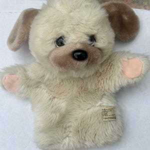 May include: A plush dog hand puppet with a cream-colored body, brown ears, and a black nose. The puppet has pink accents on its paws and a tag with text. This soft toy is designed for children.