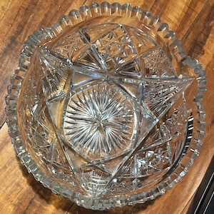 May include: Clear glass bowl with intricate starburst and geometric patterns. The bowl has a scalloped rim and a detailed design throughout. This decorative bowl is suitable for serving or display.