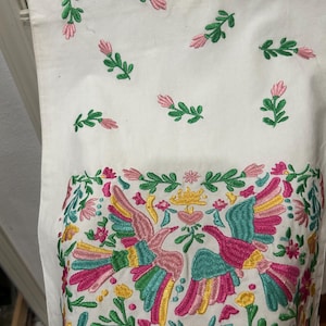 May include: White fabric with embroidered floral and bird designs. The bottom features a vibrant, colourful bird and floral pattern in pink, yellow, green, and blue. Scattered across the top are smaller embroidered pink flowers and green leaves.