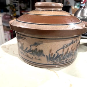 May include: A round, tan ceramic casserole dish with a matching lid. The exterior features a dark brown band around the lid and rim, with a hand-painted blue and brown brushstroke pattern depicting reeds or grass encircling the base.