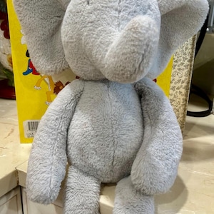 May include: A soft, gray plush elephant toy with large ears and a trunk. The elephant is sitting in front of a yellow book with illustrations. The toy is made of a fuzzy material.