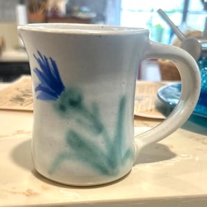 May include: A handmade ceramic mug with a white base and a blue and green floral design. The mug has a curved handle and a slightly flared top. The floral design appears to be hand-painted, adding to its unique charm.