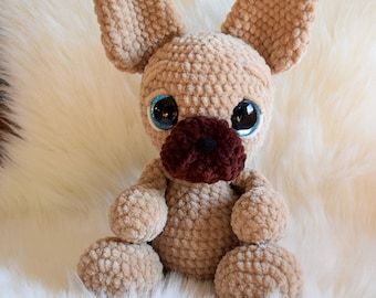 May include: A crocheted plush toy dog with brown fur, large blue eyes, and a brown nose. The dog is sitting on a white fluffy surface.
