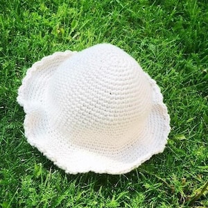 May include: A white crocheted sun hat with a scalloped brim, lying on a bed of green grass.