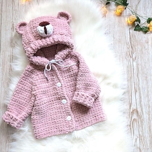 May include: A light pink, hooded baby jacket with a bear face design. The jacket has a hood with bear ears and a brown nose, white buttons, and a white drawstring. The jacket is made of a soft, textured material.