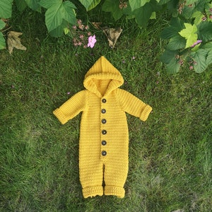 May include: A yellow crocheted baby onesie with a hood and button closure. The onesie is lying on a bed of green grass.