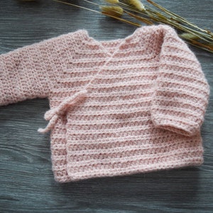 Puede incluir: Un suéter de bebé de ganchillo rosa con cierre de corbata envolvente.