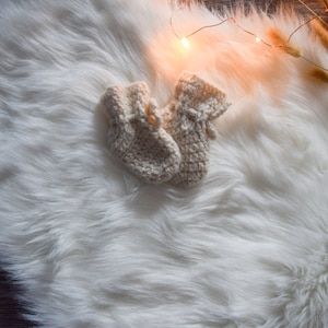 Könnte beinhalten: Zwei Paar gestrickte Babyschuhe in Cremefarbe, die auf einem weißen, flauschigen Teppich liegen. Die Schuhe sind aus weichem, strukturiertem Garn gefertigt und haben ein schlichtes Design.
