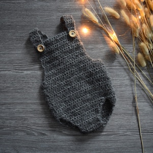 Puede incluir: Un pelele de bebé de crochet gris con botones de madera. El pelele está sobre un fondo de madera oscura.