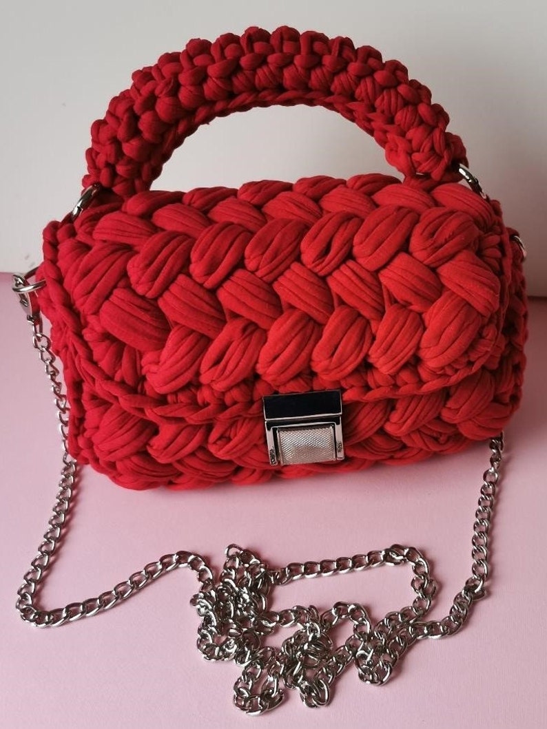 May include: A red crocheted handbag with a silver chain strap and a silver clasp. The bag has a woven texture and a top handle.