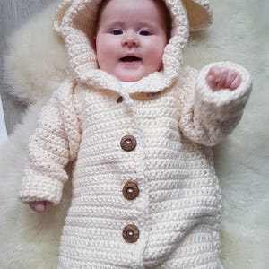 May include: A white crocheted baby onesie with a hood and bear ears. The onesie has wooden buttons down the front.