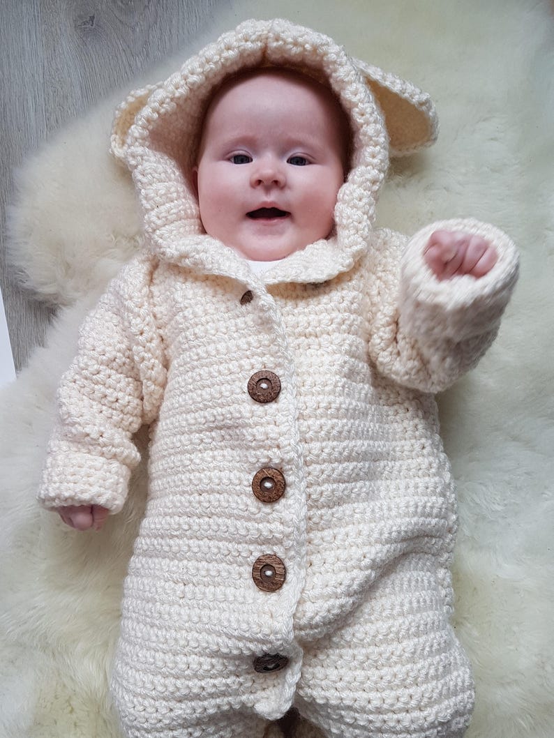 May include: A white crocheted baby onesie with a hood and bear ears. The onesie has wooden buttons down the front.