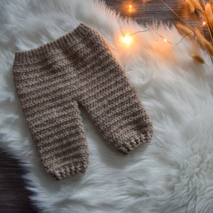 Può includere: Un paio di pantaloni per neonati in crochet beige su uno sfondo di pelliccia sintetica bianca.