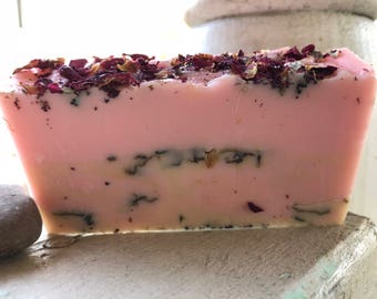 May include: A pink soap bar with dried rose petals and other botanicals on top. The soap is handmade and has a natural, earthy scent.