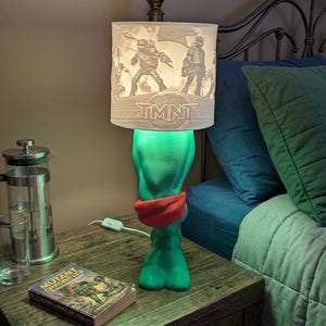 May include: A table lamp featuring a green base shaped like a character with a red mask, topped with a cylindrical white lampshade. The shade displays a graphic of four figures in action poses and the text TMNT printed in a bold, stylized font.