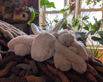 May include: A white plush toy shaped like a unicorn with a horn and wings. The unicorn is lying on a brown and gray knitted blanket.