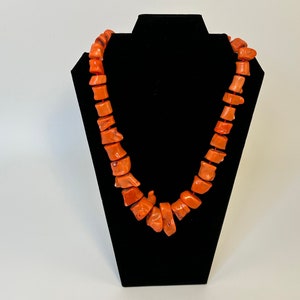 May include: A long necklace made of orange coral beads. The beads are irregular in shape and size, and they are strung together on a thin cord. The necklace is displayed on a black velvet stand.