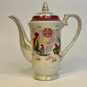 Pode incluir: Bule de porcelana branca com um design de dragão e fênix vermelho, dourado e verde. A chaleira tem uma tampa vermelha com um botão dourado e uma faixa vermelha ao redor da parte superior. A chaleira tem um esmalte branco iridescente.