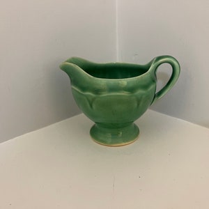 Peut inclure: Un petit pot à crème en céramique vert avec un pied et une anse. Le pot à crème a une forme légèrement arrondie et une finition lisse et brillante.