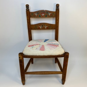 May include: A vintage wooden chair with a hand-painted floral design on the backrest. The seat features a patchwork quilt in red, white, and blue fabrics. The chair has a rustic, farmhouse aesthetic.