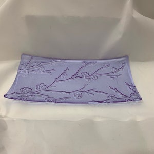 May include: A rectangular, lavender-colored glass dish with a floral branch design. The branches and flowers are embossed on the surface. The dish has slightly curved sides and is suitable for serving or display.